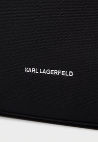 Černá texturovaná látková taška s logem „KARL LAGERFELD“ v bílé barvě výrazně umístěným na boku. Čistý design s minimálními akcenty.