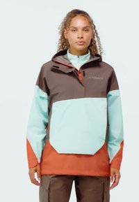 Žena s kudrnatými vlasy nosící venkovní bundu ve více barvách, s hnědými, mátově zelenými a oranžovými částmi, stojící na jednobarevném pozadí.