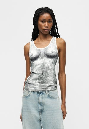 Ženska z dolgimi kitkami, oblečena v brezrokavnik z natančnim črno-belim umetniškim tiskom na trup in svetlo modrimi jeans hlačami.