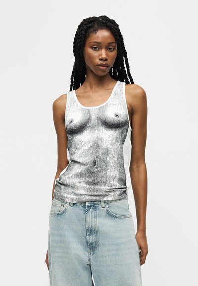 Femme aux longues tresses portant un débardeur sans manches avec une impression artistique en noir et blanc détaillée sur le torse et un jean en denim bleu clair.