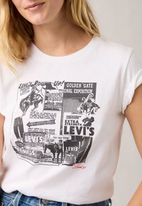 Λευκό βαμβακερό μπλουζάκι με μαύρα τυπωμένα γραφικά από vintage διαφημίσεις της Levi's, με κόκκινες λεπτομέρειες και μανίκια που είναι γυρισμένα.
