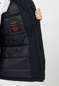 Schwarze Jacke mit einem gesteppten Innenfutter, ausgestattet mit einem orangefarbenen Logo-Patch mit der Aufschrift "BOMBERS". Mit Reißverschluss und Druckknöpfen vorne.