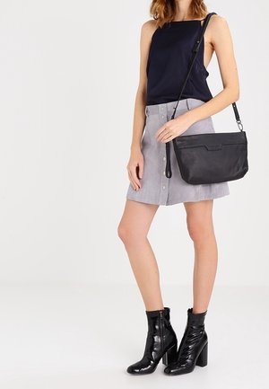 Czarna skórzana torebka na ramię z klapką, zestawiona z granatową bezrękawną bluzką, szarą spódnicą na guziki z przodu oraz błyszczącymi czarnymi botkami za kostkę.