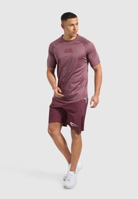 Bordeauxrotes Sport-T-Shirt mit kurzen Ärmeln und Logo auf der Vorderseite; kombiniert mit passenden Shorts, die über eine Seitentasche und weiße Akzentdetails verfügen.
