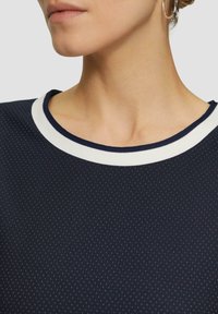 Haut bleu marine avec un col blanc; tissu texturé présentant de petites rayures verticales régulièrement espacées. Gros plan du décolleté.
