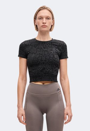 Kvinde med kort blondt hår iført sort mønstret cropped t-shirt og høj-taljede taupe leggings med Adidas-logo, står mod en ensartet baggrund.