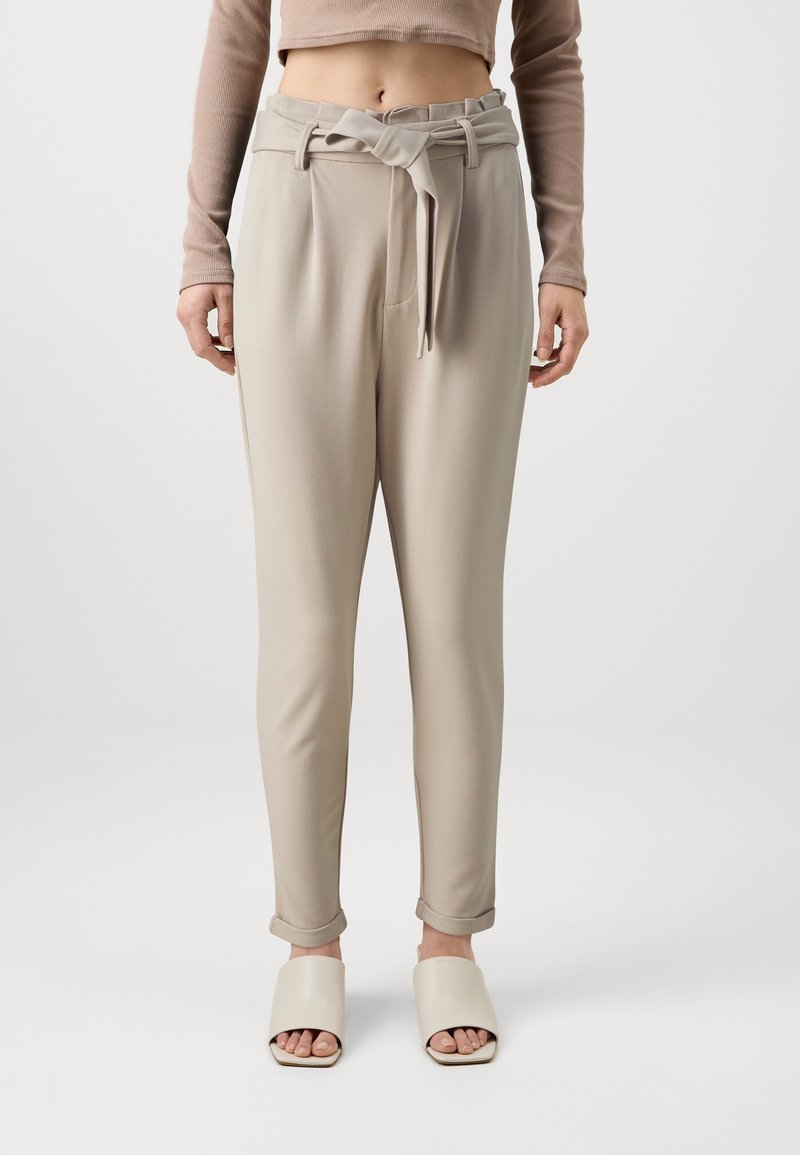 Femme portant un pantalon beige taille haute style paperbag avec une ceinture nouée et des ourlets roulottés, avec des sandales crème à bout ouvert.