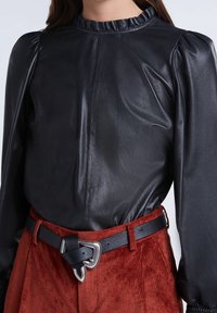 Svart läderblus med volangkrage och puffärmar, tillsammans med rostfärgade manchesterbyxor och ett svart bälte med en silverspänne.