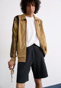 Dzeltena zip-up jaka ar melniem akcentiem un trim svītrām; baltā T-kreklā apakšā; melnas šortas; tur rokās brilles.