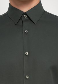 Mørkegrønn skjorte med knapper og klassisk krage, med en glatt tekstur og grå knapper langs fronten for feste.