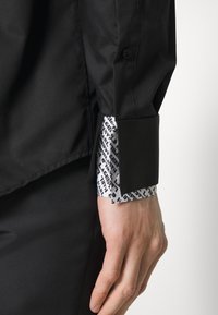 Černá košile s elegantním designem, která má kontrastní bílé manžety s černým textovým vzorem. Manžety na knoflíky a hladká textilie jsou zmíněny.