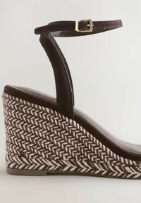 Sandalo con zeppa marrone e suola in fibra naturale intrecciata. Presenta un cinturino alla caviglia marrone con fibbia dorata e dettagli in pelle liscia sulla parte superiore.
