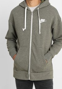 Olivgrüne Zip-Hoodie mit einem weißen Nike-Logo, Kordelzügen, Fronttaschen und gerippten Bündchen. Weicher, texturierter Stoff.