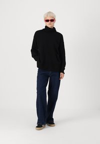 Črna oversize turtleneck pulover z rebrasto teksturo, v kombinaciji s temno modrimi širokimi hlačami in črnimi čevlji s svetlim podplatom.