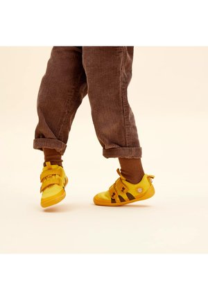 Kind trägt braune Cordhose und gelbe Klettverschluss-Sneaker, steht auf einem Bein vor hellem beigefarbenem Hintergrund.