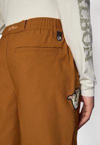 Pantalon marron texturé avec une taille élastique, doté d'une poche arrière et d'un motif brodé coloré sur le côté.