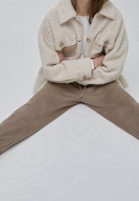 Person med en fluffig beige jacka och beiga byxor, sittande i skräddarställning på en ljus bakgrund med armarna i kors, som förmedlar en avslappnad pose.