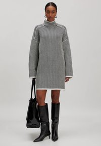 Robe en tricot oversize avec des rayures verticales noires et blanches, col montant, épaules tombantes et texture côtelée. Portée avec des bottes noires jusqu'aux genoux.