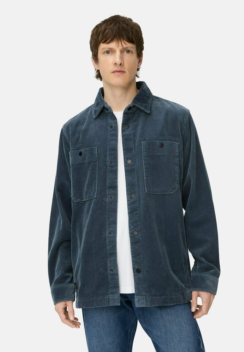 Ung mand iført en blå kordfløjl skjorte med knapper over en hvid T-shirt og blå jeans, mod en simpel lys baggrund.