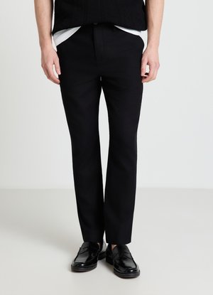 Uomo con pantaloni neri su misura, mocassini neri e un top nero con orlo bianco, in piedi davanti a uno sfondo chiaro e uniforme.