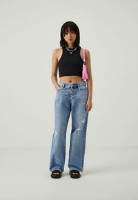 Mulher vestindo um top cropped preto, jeans azul desgastado de cintura alta com botões, sandálias pretas, colares coloridos e carregando uma bolsa de ombro rosa.