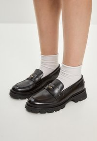 Musta nahka loaferit tukevalla kumipohjalla, joissa on sileä pinta, koristeellinen hihna ja pieni kultainen yksityiskohta.