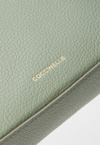 Green textured leather bag with gold embossed "Coccinelle" logo. Features a smooth, slightly glossy finish and contrasting stitching.