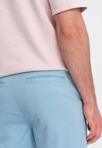 Světle růžová bavlněná polokošile v kombinaci se světle modrými kraťasy, které mají zadní kapsu a hladkou texturu. Viditelný šev na košili.