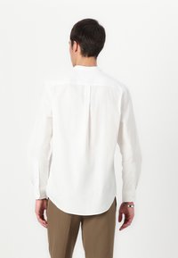 Camicia bianca a maniche lunghe con orlo arrotondato, piega posteriore e polsini con bottoni; il tessuto sembra leggero e morbido. Abbinata a pantaloni marroni.