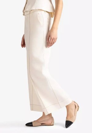 Person wearing beige wide-leg pants with visible stitching and beige flats with black toe caps, standing sideways on a white background.