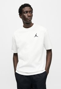 Λευκή βαμβακερή κοντομάνικη μπλούζα με στρογγυλή λαιμόκοψη, με μαύρο λογότυπο Jumpman στο αριστερό στήθος, συνδυασμένη με σκούρο σορτς.