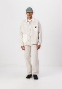 Striped cotton jacket with a cream body and light beige stripes, paired with matching striped pants. Black shoes and a green cap complete the look.