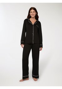 Donna sorridente, in piedi a piedi nudi, indossa un pigiama nero a maniche lunghe con bottoni, con piping bianco su colletto, polsini, tasca e orli dei pantaloni.