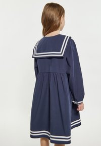 Marineblaues Kleid mit einem Matrosenkragen, der weiße Streifen aufweist, langen Ärmeln und einer gerafften Taille, die am Saum ausgestellt ist.