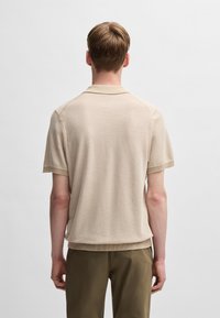 Polo din tricot bej cu mâneci scurte, având un guler clasic și o textură subtilă striată, asortat cu pantaloni maro deschis.