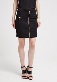 Jupe noire ajustée avec des fermetures éclair dorées à l'avant et deux poches latérales. Texture lisse, longueur au-dessus du genou, accompagnée de talons noirs à lanières.