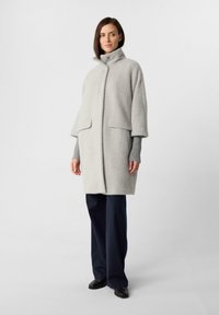 Donna in piedi con un cappotto grigio chiaro a colletto alto, maniche a costine grigie, pantaloni neri a gamba alta e scarpe nere su uno sfondo neutro.