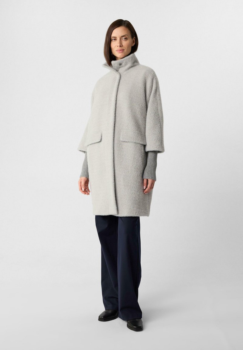 Donna in piedi con un cappotto grigio chiaro a colletto alto, maniche a costine grigie, pantaloni neri a gamba alta e scarpe nere su uno sfondo neutro.