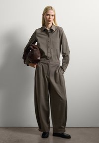Joven mujer con largo cabello rubio, vistiendo una camisa marrón holgada y pantalones de pierna ancha, sosteniendo un gran bolso de cuero marrón.