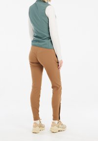 Bezramienna chabrowa kamizelka z elastycznego materiału, sparowana z dopasowanymi brązowymi spodniami z bocznymi zamkami; noszona z masywnymi beżowymi sneakersami.