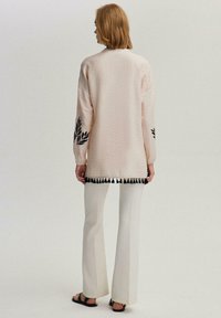 Cardigan rosa chiaro in tessuto testurizzato, con ricami neri a foglia sulle maniche e bordo a frange nere in fondo. Abbinato a pantaloni bianchi a zampa.