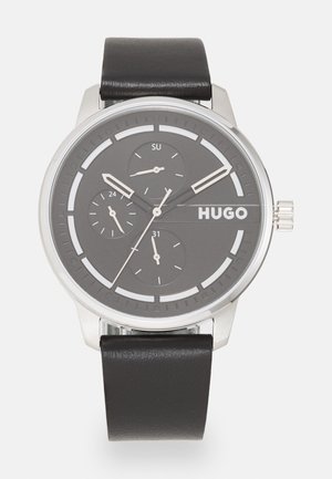 Montre analogique argentée avec un cadran gris rond, trois sous-cadrans, un bracelet en cuir noir et des marquages blancs ; le nom de la marque "HUGO" affiché en évidence.