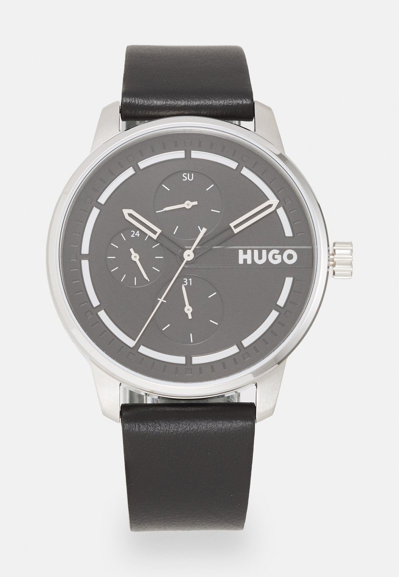 Silberne Analog-Uhr mit einem runden grauen Zifferblatt, drei Unterzifferblättern, schwarzem Lederarmband und weißen Markierungen; der Markenname "HUGO" ist auffällig dargestellt.