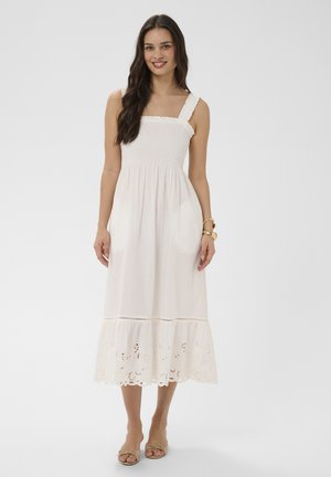 Kobieta o długich ciemnych włosach, ubrana w białą, bezrękawną sukienkę midi z marszczonym gorszem i haftowanym dołem, beżowe sandały oraz bransoletki, uśmiechająca się.