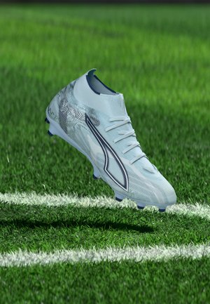 Weißer und silberner Fußballschuh mit schwarzem Logo schwebt über grünem Gras nahe einer weißen Spielfeldlinie.