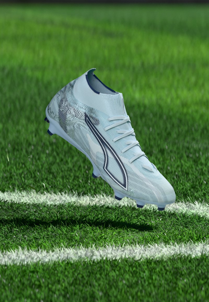 Beli in srebrni nogometni čevelj s črnim logotipom, ki lebdi nad zeleno travo ob belem mejniku na igrišču.