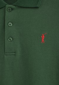 Grünes Polohemd aus strukturiertem Stoff, mit drei dunklen Knöpfen und einer kleinen roten, gestickten Figur, die einen Golfschläger hält, auf der linken Seite.