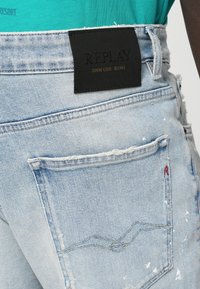 Ανοιχτό μπλε τζιν παντελόνι με φθαρμένες λεπτομέρειες, διαθέτει μαύρη ετικέτα που γράφει "REPLAY" στη μέση και διακοσμημένες πίσω τσέπες.