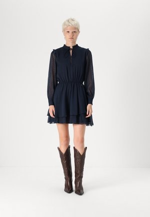 Navy blue dress with long sheer sleeves, ruffled hem, and a front tie detail, paired with tall, dark brown cowboy boots with embossed patterns.