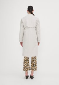 Lys grå trenchcoat med strukturerede skuldre, der har en bagklap. Pairing med mønstrede vide bukser i gul, sort og hvid.